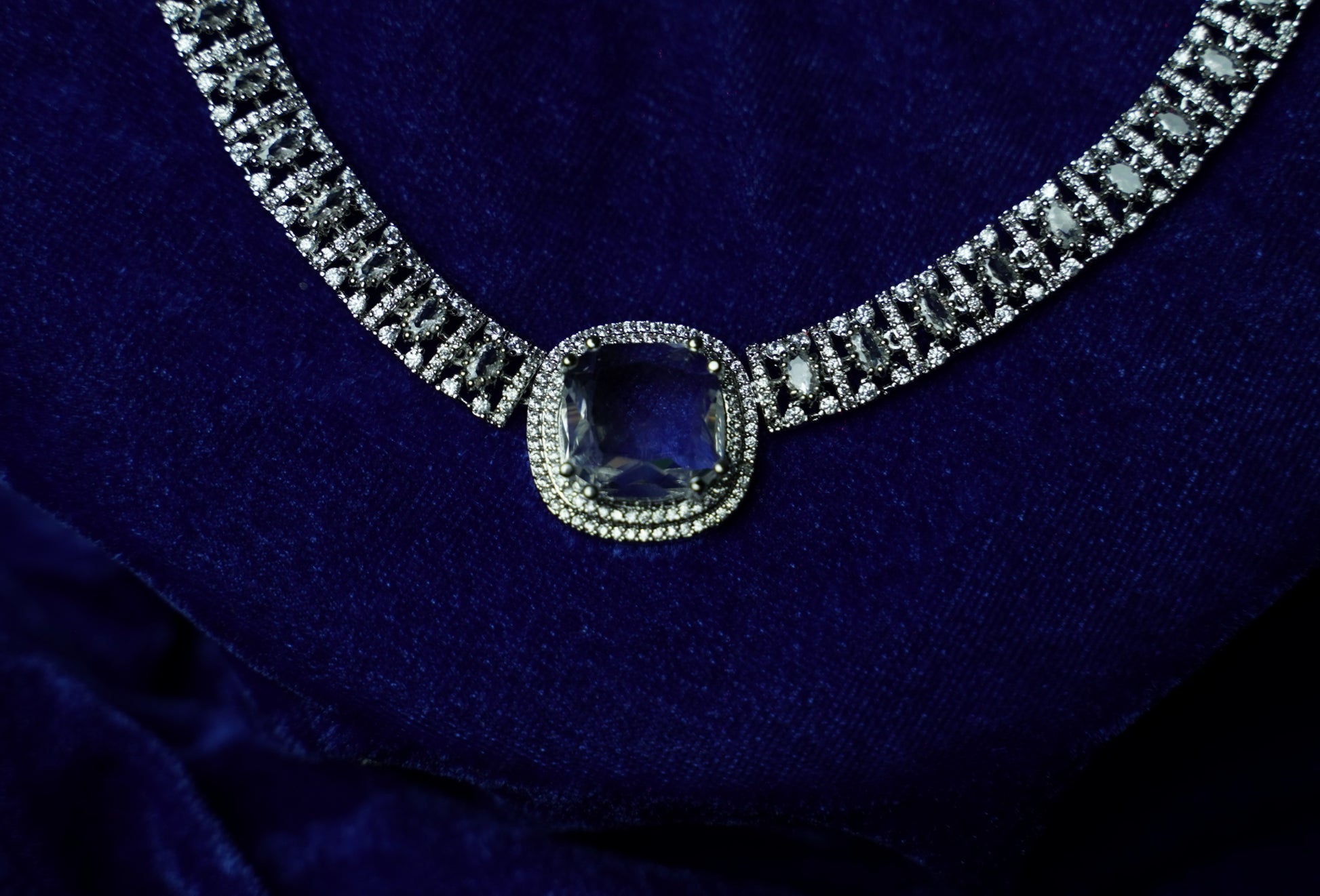 A close-up of a diamond-studded necklace with a central cushion-cut blue gemstone, displayed on a deep blue velvet surface.