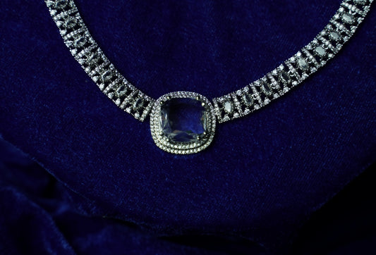 A close-up of a diamond-studded necklace with a central cushion-cut blue gemstone, displayed on a deep blue velvet surface.