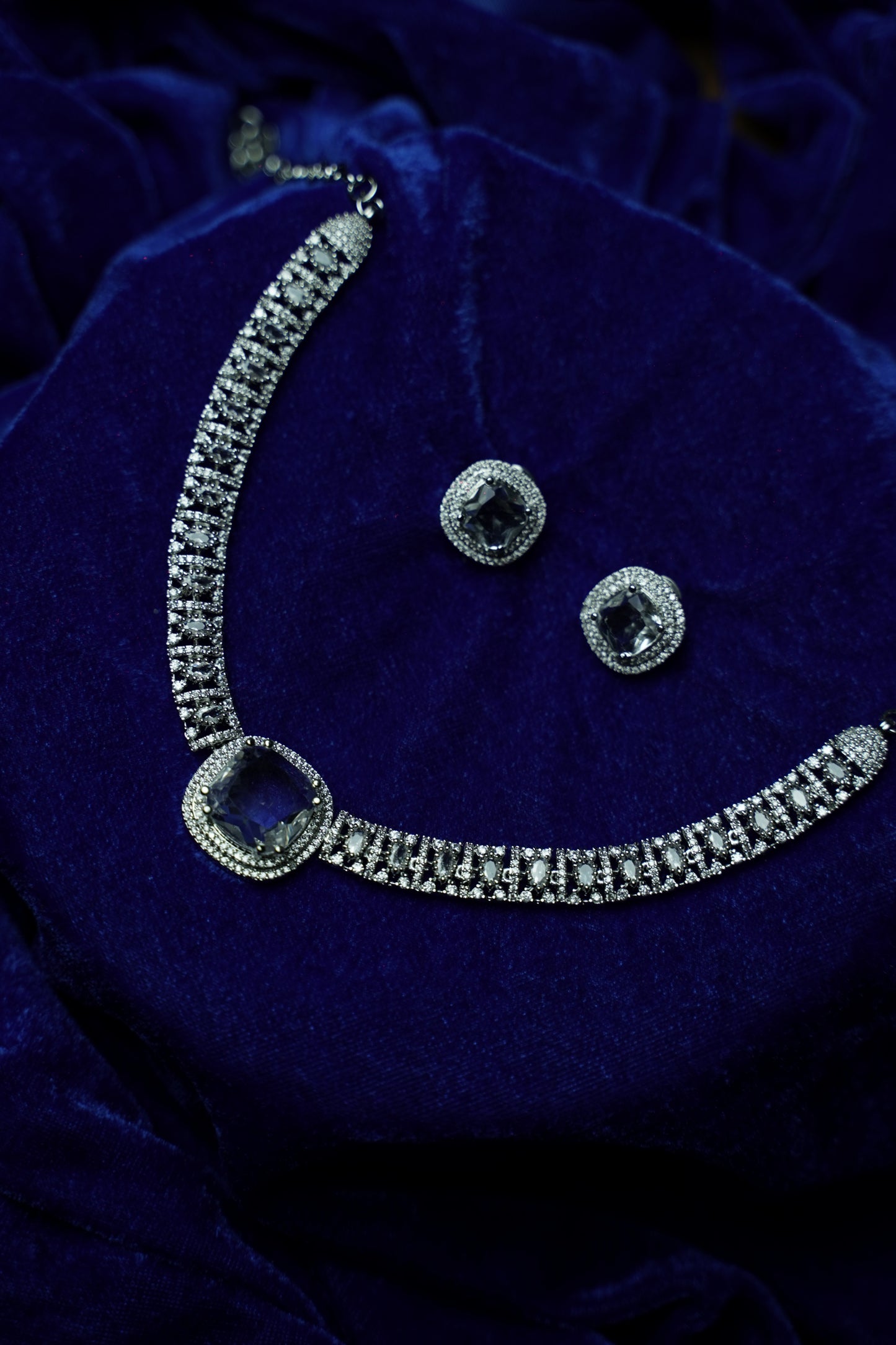 Silver-toned jewelry set featuring a necklace with a square-cut blue center stone and matching stud earrings, displayed on a deep blue velvet fabric background.
