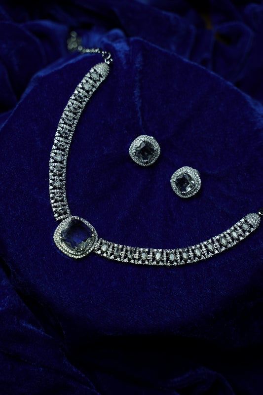 Silver-toned jewelry set featuring a necklace with a square-cut blue center stone and matching stud earrings, displayed on a deep blue velvet fabric background.