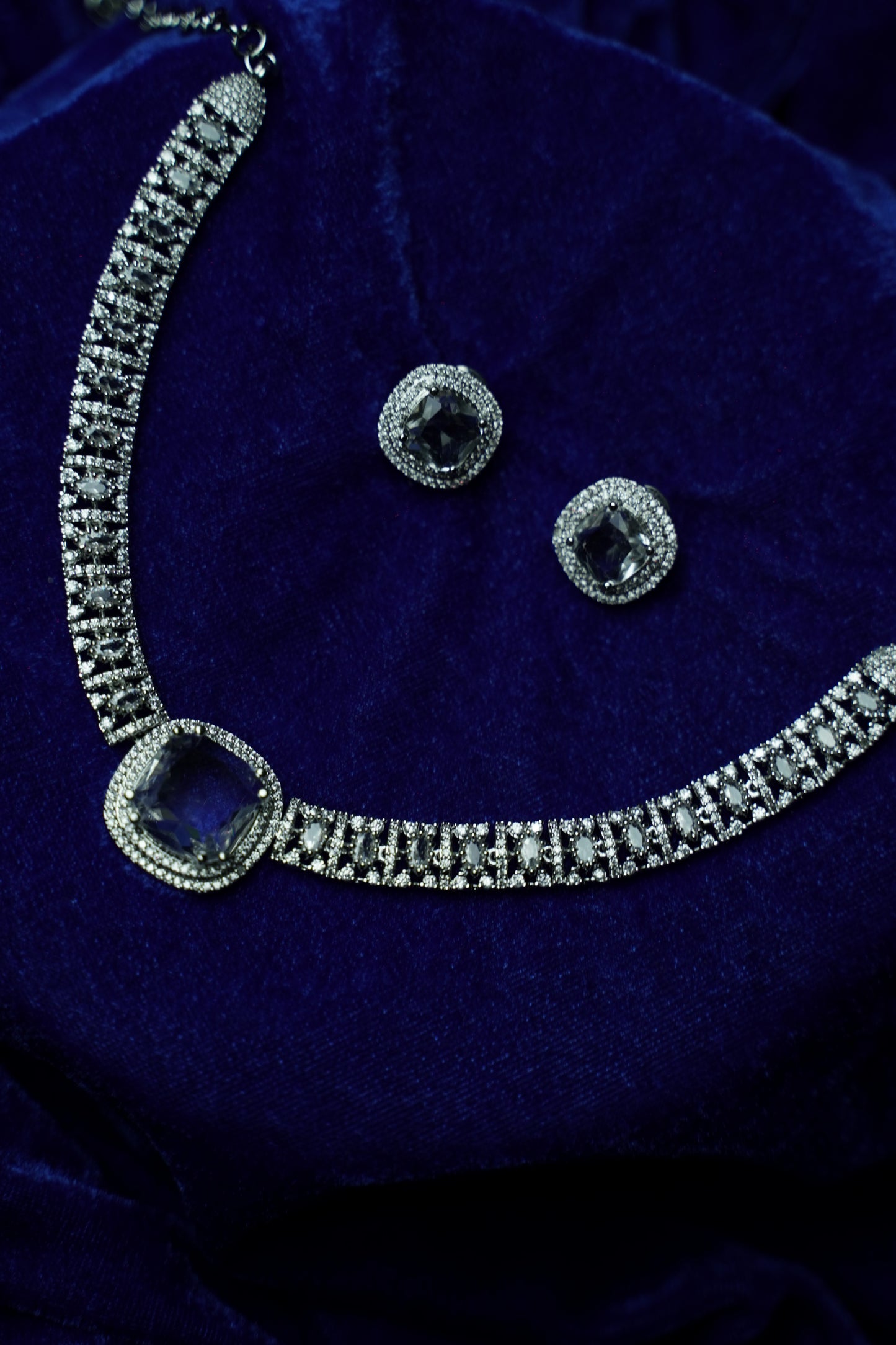 Silver-toned jewelry set featuring a necklace and matching earrings with clear stones and a large dark blue gemstone, displayed on a deep blue velvet surface.