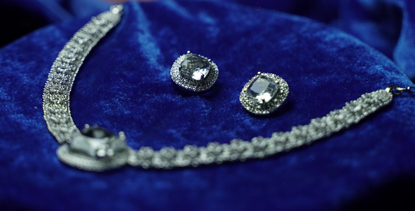 A silver-toned jewelry set featuring a necklace and matching stud earrings with large clear gemstones, displayed on a deep blue velvet fabric background.