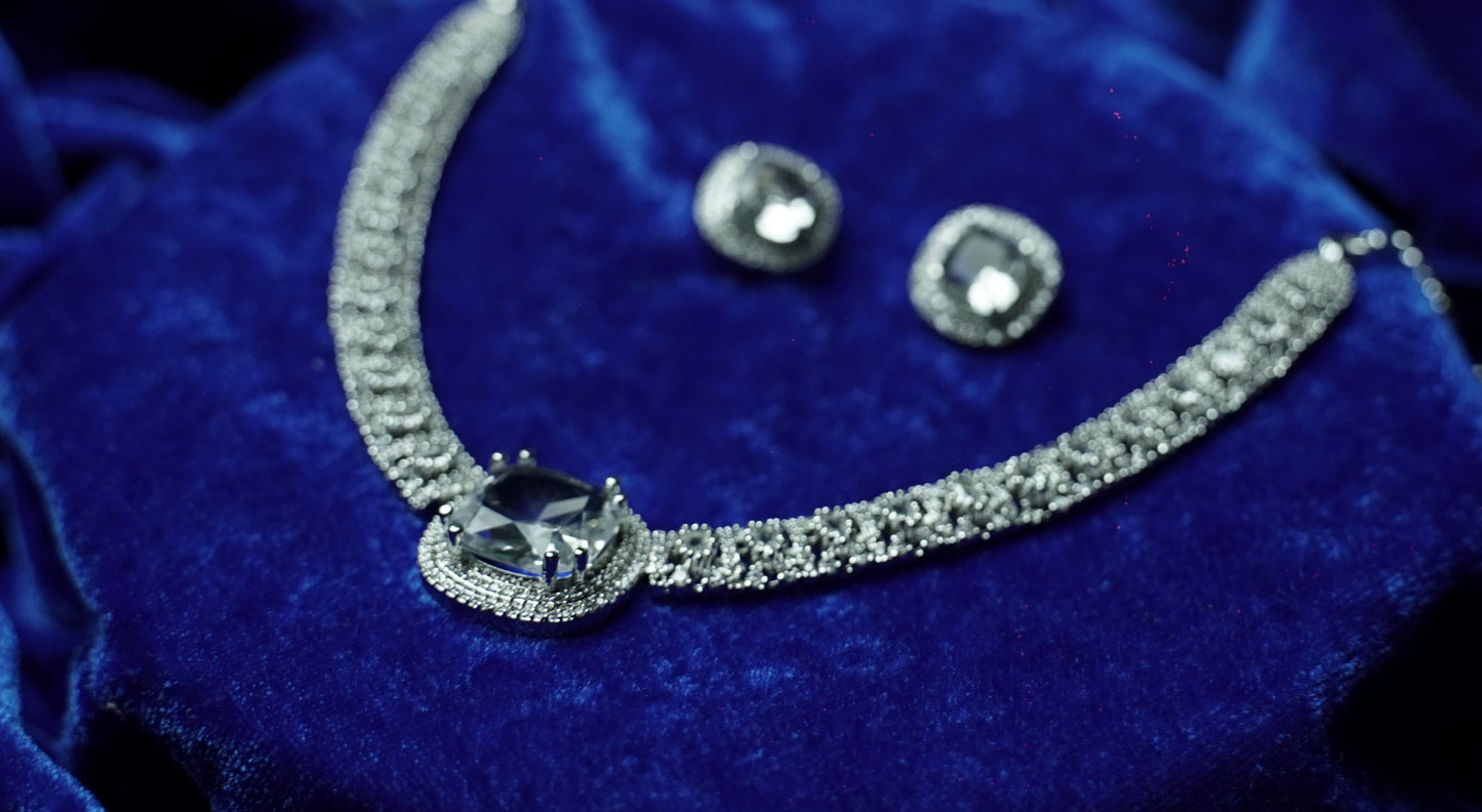 A silver-toned jewelry set featuring a necklace and matching earrings with clear gemstones, displayed on a deep blue velvet fabric background.