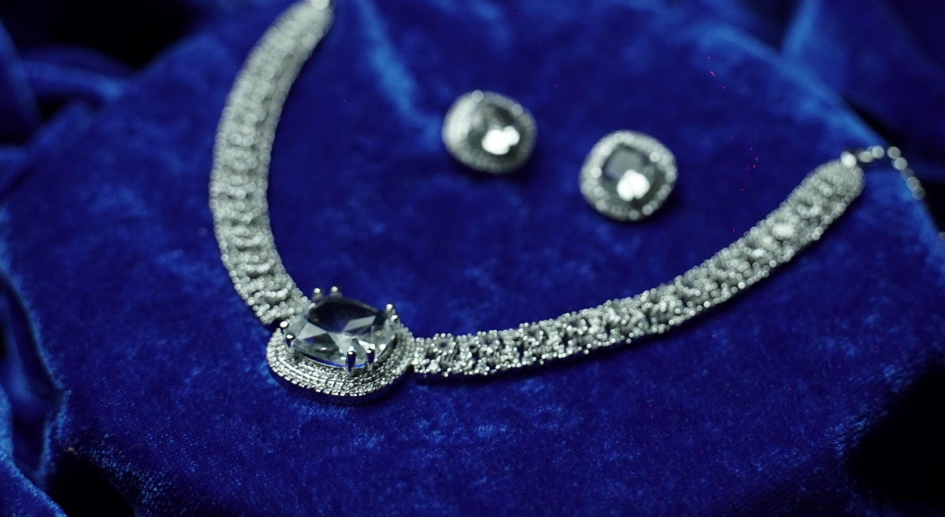A silver-toned jewelry set featuring a necklace and matching earrings with clear gemstones, displayed on a deep blue velvet fabric background.
