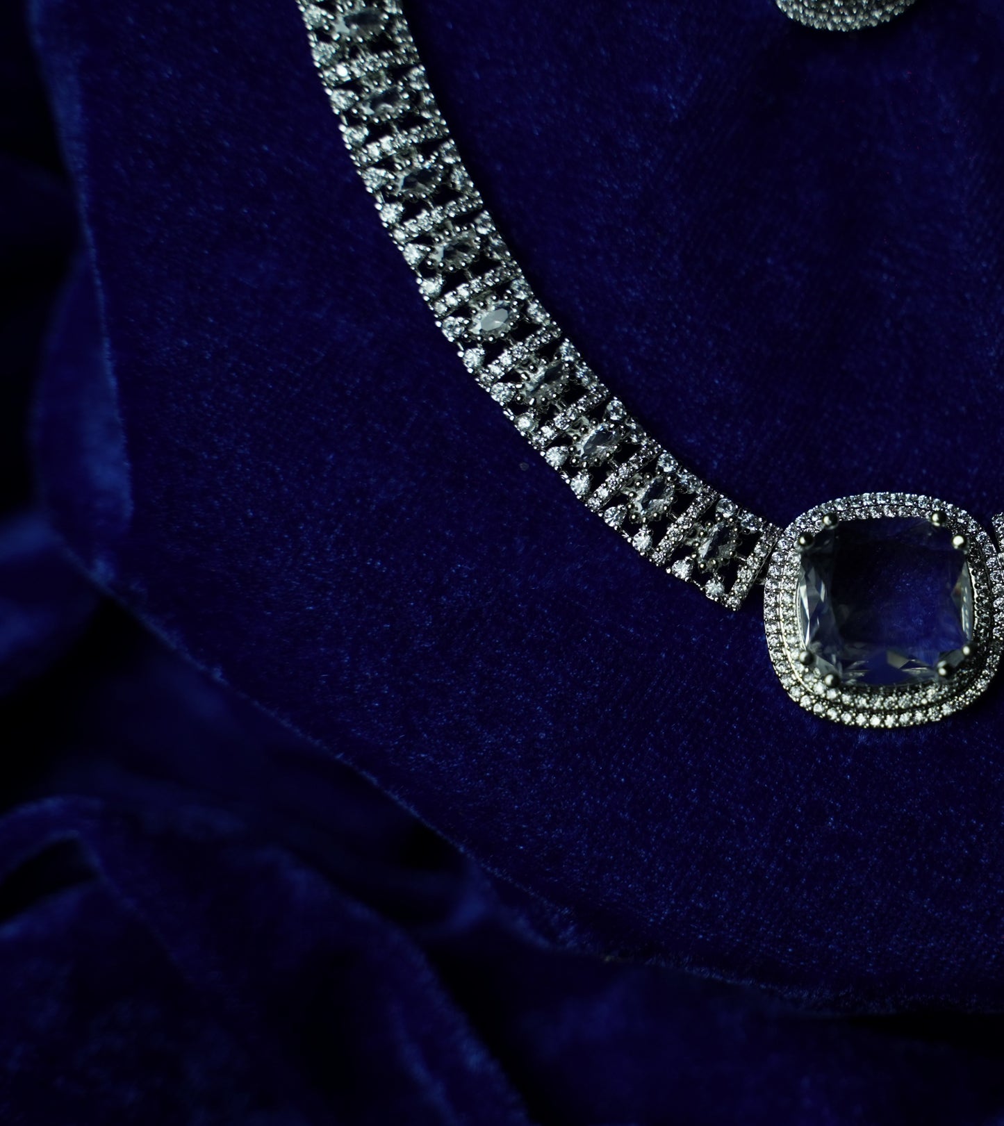 A close-up of a silver-toned necklace with a large cushion-cut blue gemstone pendant, displayed on a deep blue velvet surface.