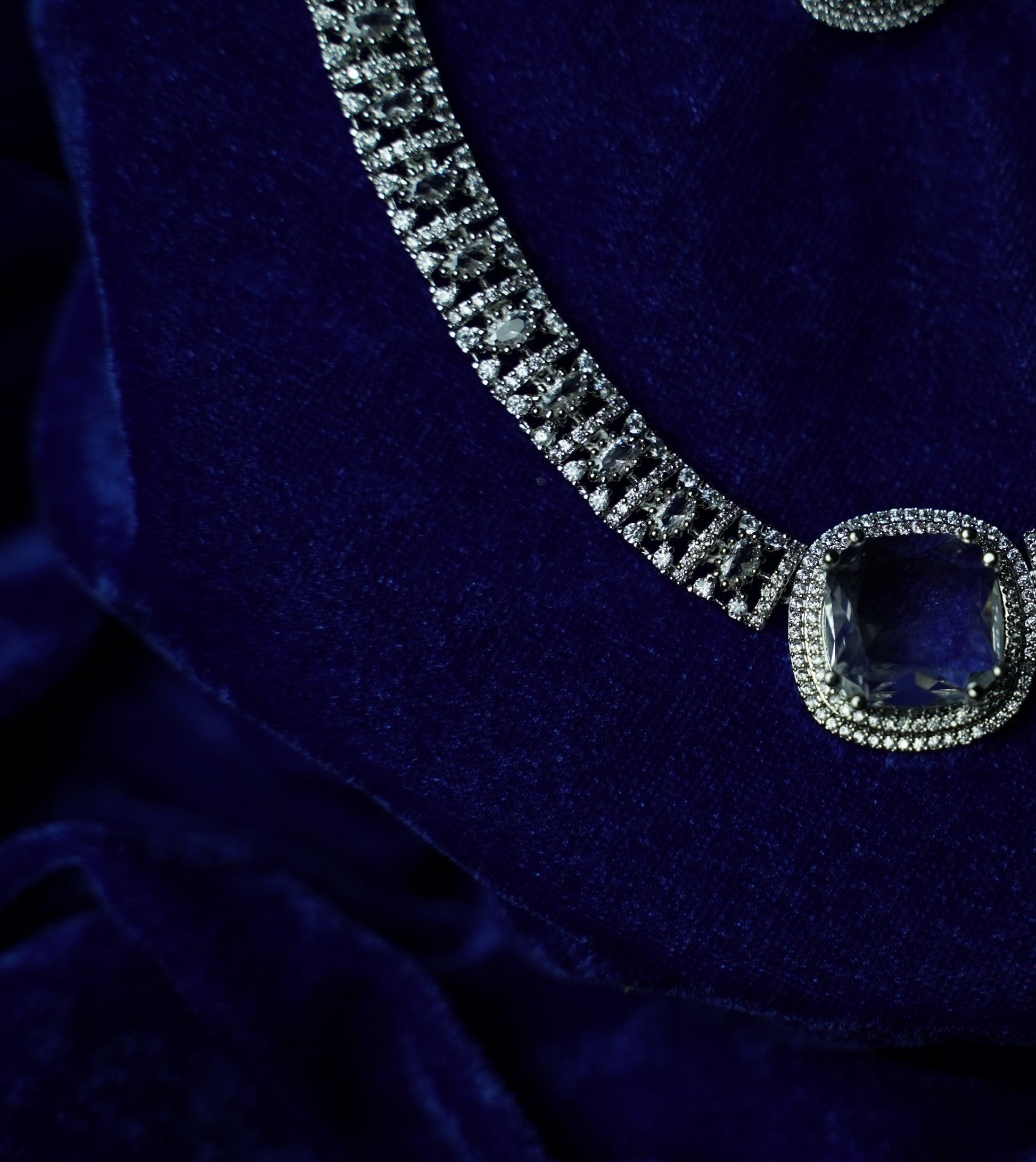 A close-up of a silver-toned necklace with a large cushion-cut blue gemstone pendant, displayed on a deep blue velvet surface.